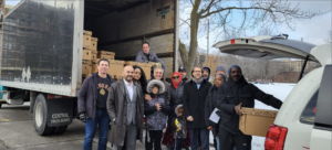 Group of people participating in a community charity event, standing near a truck and an open car trunk filled with boxes.